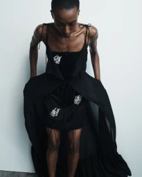 Amelie Nsengiyumva wearing an embroidered black mini dress with a dramatic sheer black overlay against an off-white background.