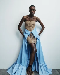 Model in a strapless beige corset top and light blue flared skirt against a white studio background for Hellessy Resort 25