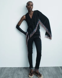 Amelie Nsengiyumva poses in a black draped fringe top, slim trousers, and platform sandals against a white wall for Hellessy Resort 25.
