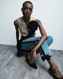 Model Amelie Nsengiyumva seated wearing a plaid vest with black fabric rosette, denim jeans with bow hems, and black shoes.