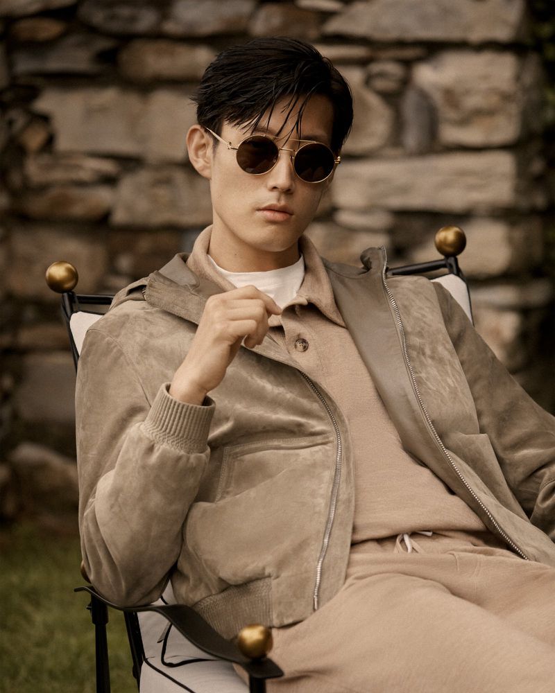 Man in sunglasses sits outdoors in a chair, wearing a tan jacket, sweater, and pants, posing against a stone wall and green background.