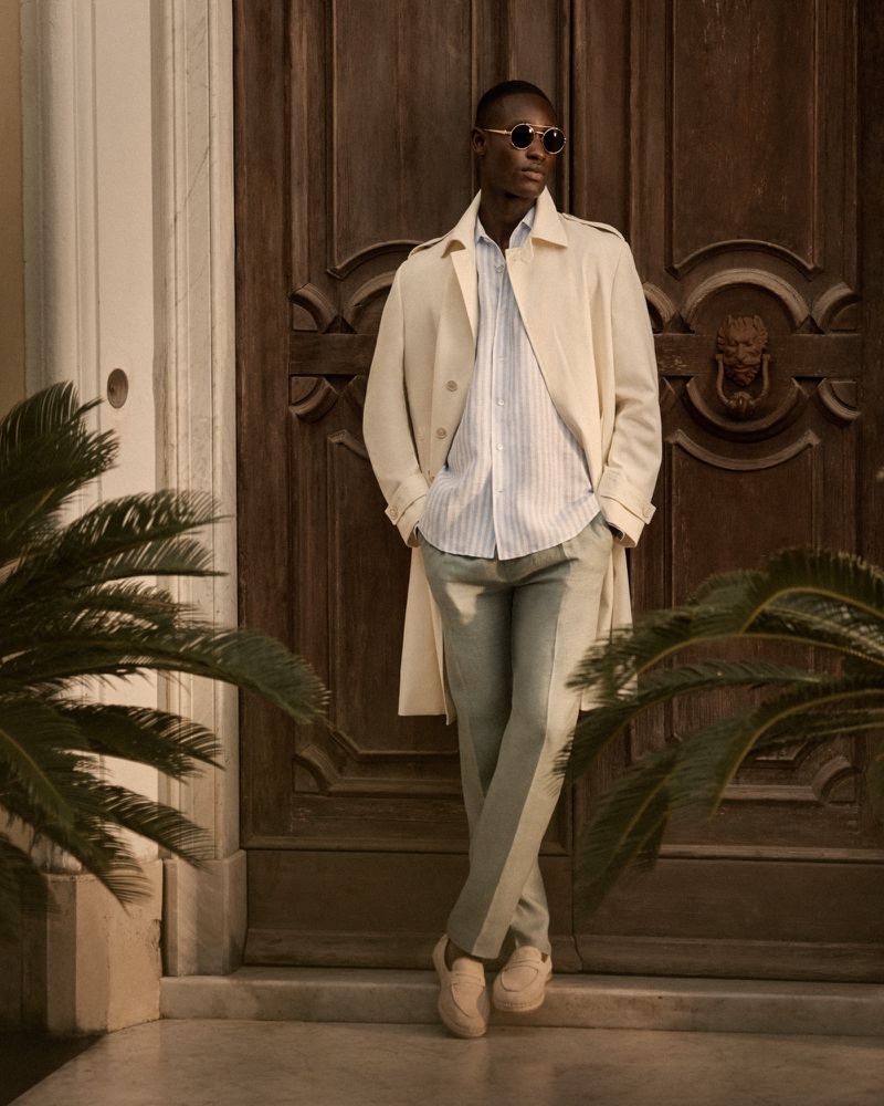 An African American man leans against a wooden door in a light-colored trench coat, blue shirt, and grey pants, standing in front of a building.