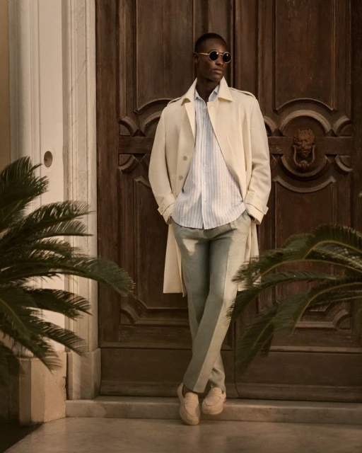 An African American man leans against a wooden door in a light-colored trench coat, blue shirt, and grey pants, standing in front of a building.