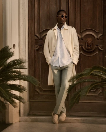 An African American man leans against a wooden door in a light-colored trench coat, blue shirt, and grey pants, standing in front of a building.
