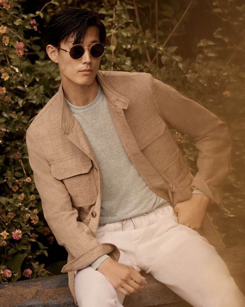 A stylish adult male sits in a tan jacket and round sunglasses outdoors with a backdrop of green foliage and small pink flowers.