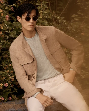 A stylish adult male sits in a tan jacket and round sunglasses outdoors with a backdrop of green foliage and small pink flowers.