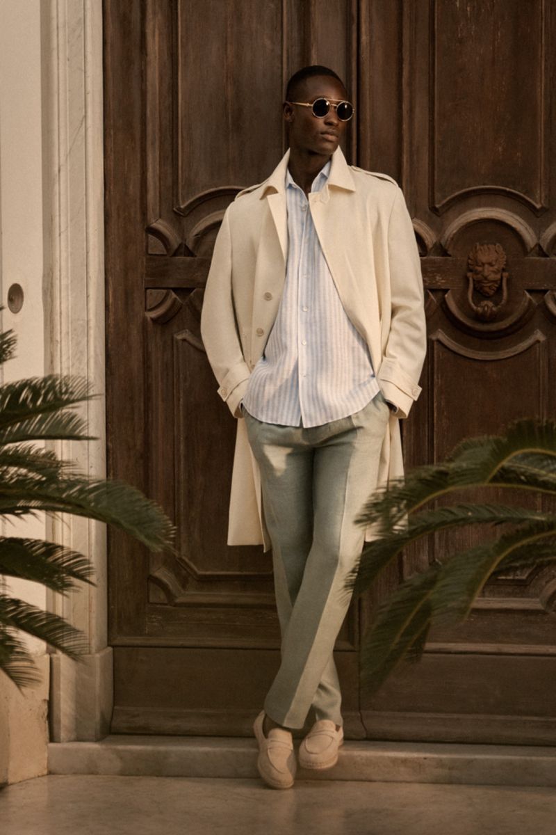 Man in trench coat and sunglasses standing in front of a wooden door, surrounded by green plants. Fashion, style, and outdoor shot.