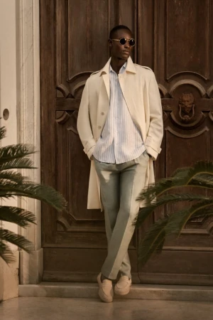 Man in trench coat and sunglasses standing in front of a wooden door, surrounded by green plants. Fashion, style, and outdoor shot.