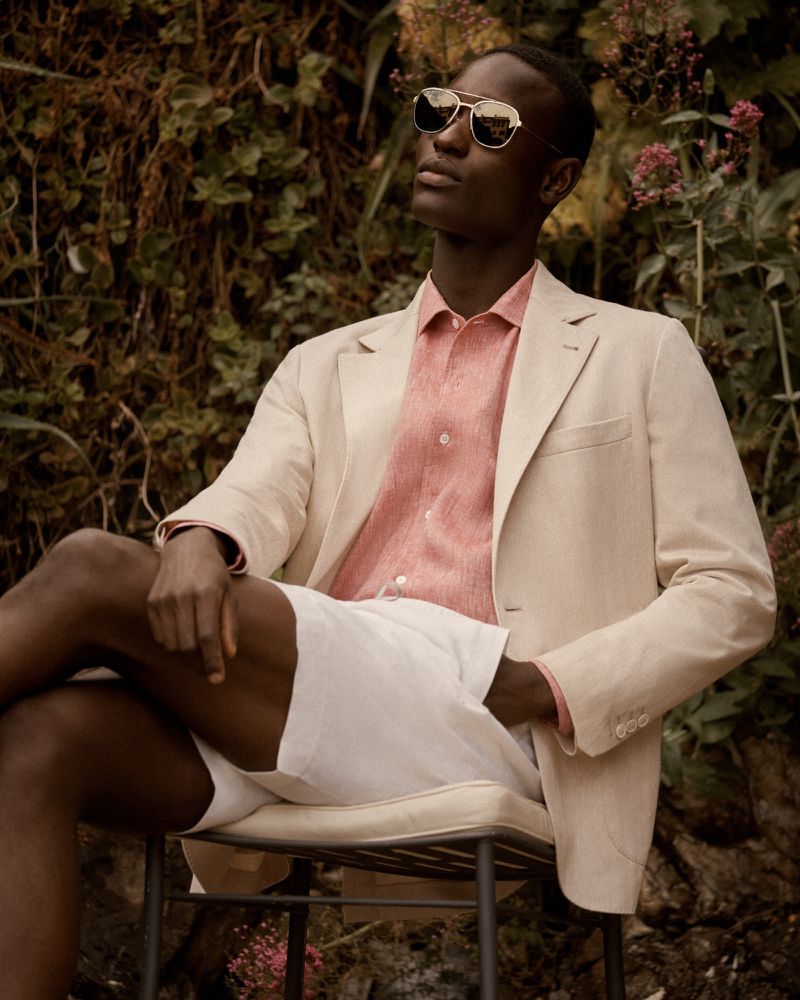 A well-dressed Black man with sunglasses sits outdoors wearing a cream blazer, coral shirt, and white shorts, looking stylish in natural light.