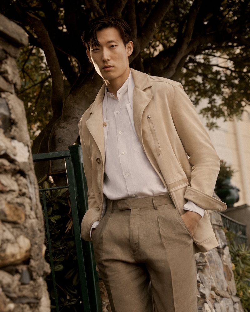 Portrait of an Asian man in beige jacket and pants standing outdoors in front of stone wall with a tree in the background.