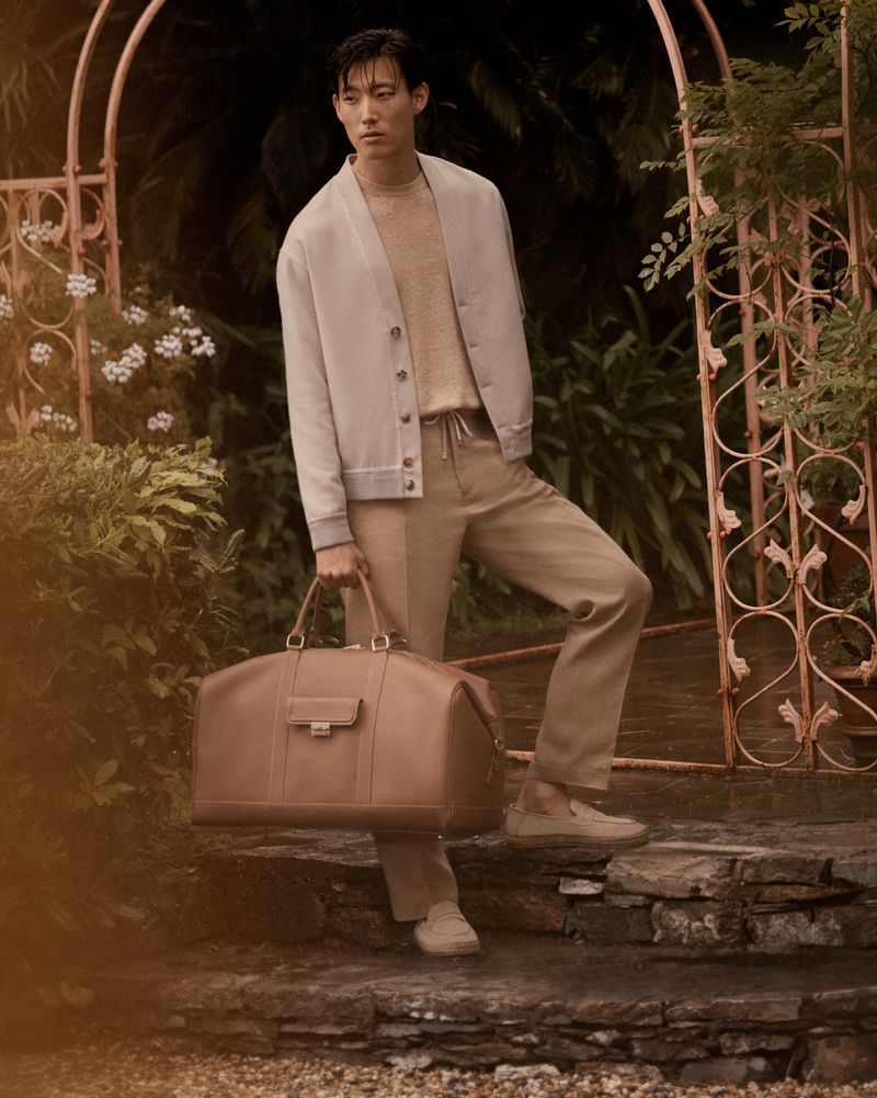 An Asian male model poses on stone steps, holding a leather duffel bag while wearing a cardigan and tan trousers in a lush garden setting.