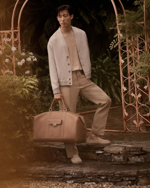 An Asian male model poses on stone steps, holding a leather duffel bag while wearing a cardigan and tan trousers in a lush garden setting.
