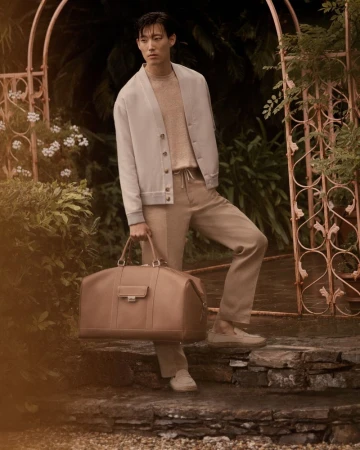 An Asian male model poses on stone steps, holding a leather duffel bag while wearing a cardigan and tan trousers in a lush garden setting.