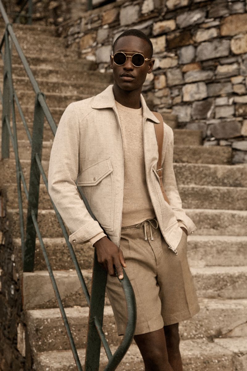Stylish dark-skinned man poses on stone steps in round sunglasses, jacket, beige shorts and sweater for a fashion editorial outdoors.