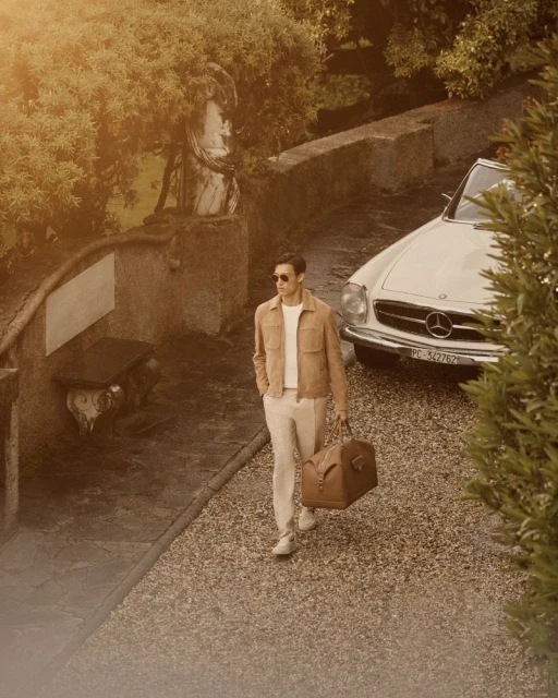 Stylish man walks along a stone path carrying a leather duffel bag next to a classic car in an outdoor, European garden setting.
