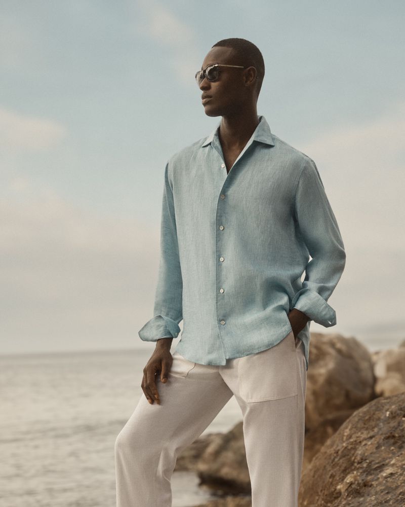A stylish Black male model wearing sunglasses and a turquoise linen shirt poses outdoors against a rocky shoreline and sky.