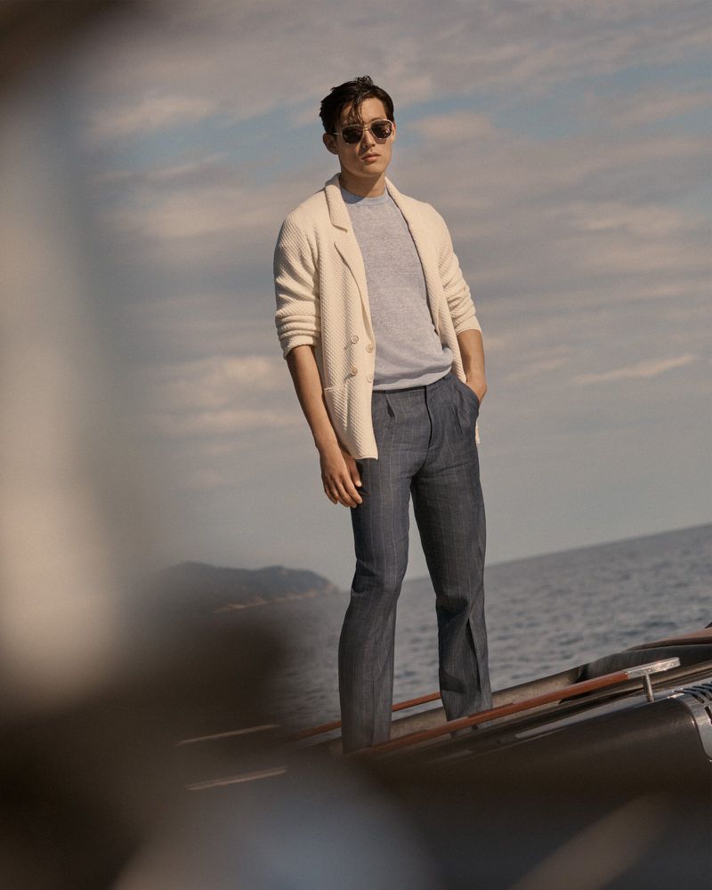 Man in sunglasses posing on a boat with the ocean and cloudy sky in the background, wearing a beige jacket, gray shirt, and gray pants.