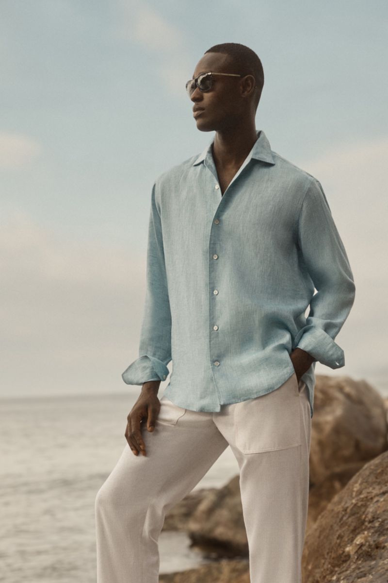 A man wearing sunglasses and a light blue linen shirt and beige pants stands on rocks near the ocean under a cloudy sky.