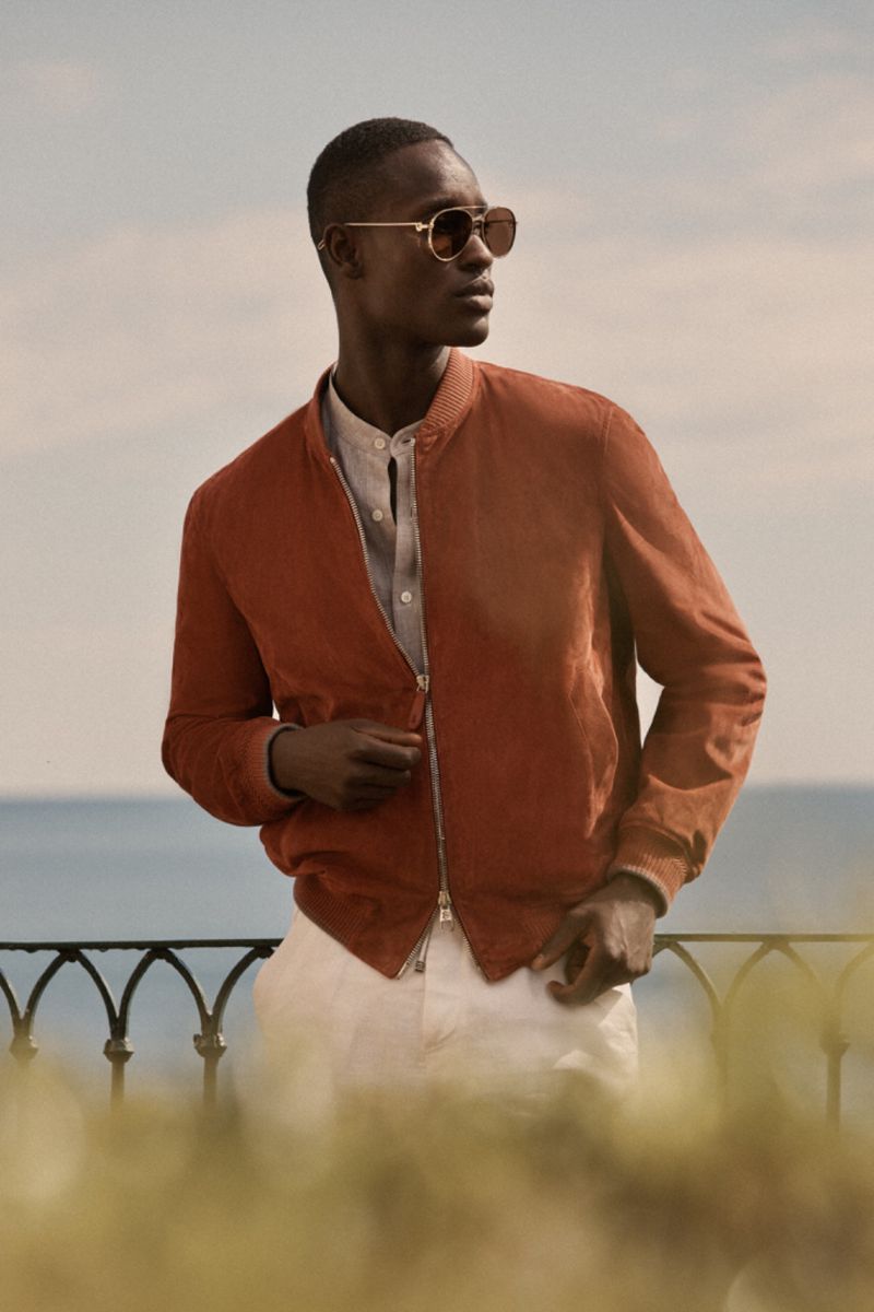 Man in rust-colored suede jacket, round sunglasses, and white pants outdoors with blue sky and ocean backdrop in medium portrait shot.