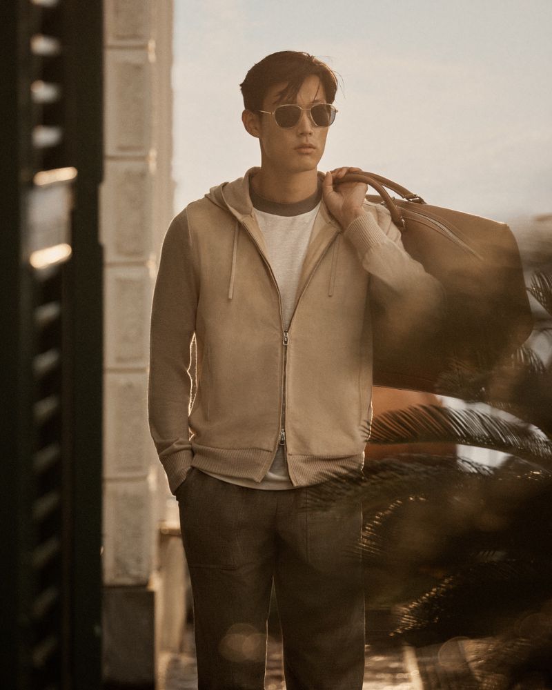 Man in a hoodie and sunglasses carries a duffel bag outside, framed by a neutral background, showcasing contemporary fashion and travel.