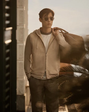 Man in a hoodie and sunglasses carries a duffel bag outside, framed by a neutral background, showcasing contemporary fashion and travel.
