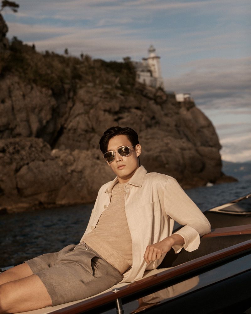 Asian man in sunglasses poses on a boat with a lighthouse and cliffs in the background. Shot with warm lighting in daylight.