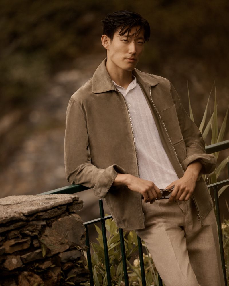 Portrait of an adult Asian man with dark hair wearing a collared shirt and a suede jacket, leaning against a railing in an outdoor setting.