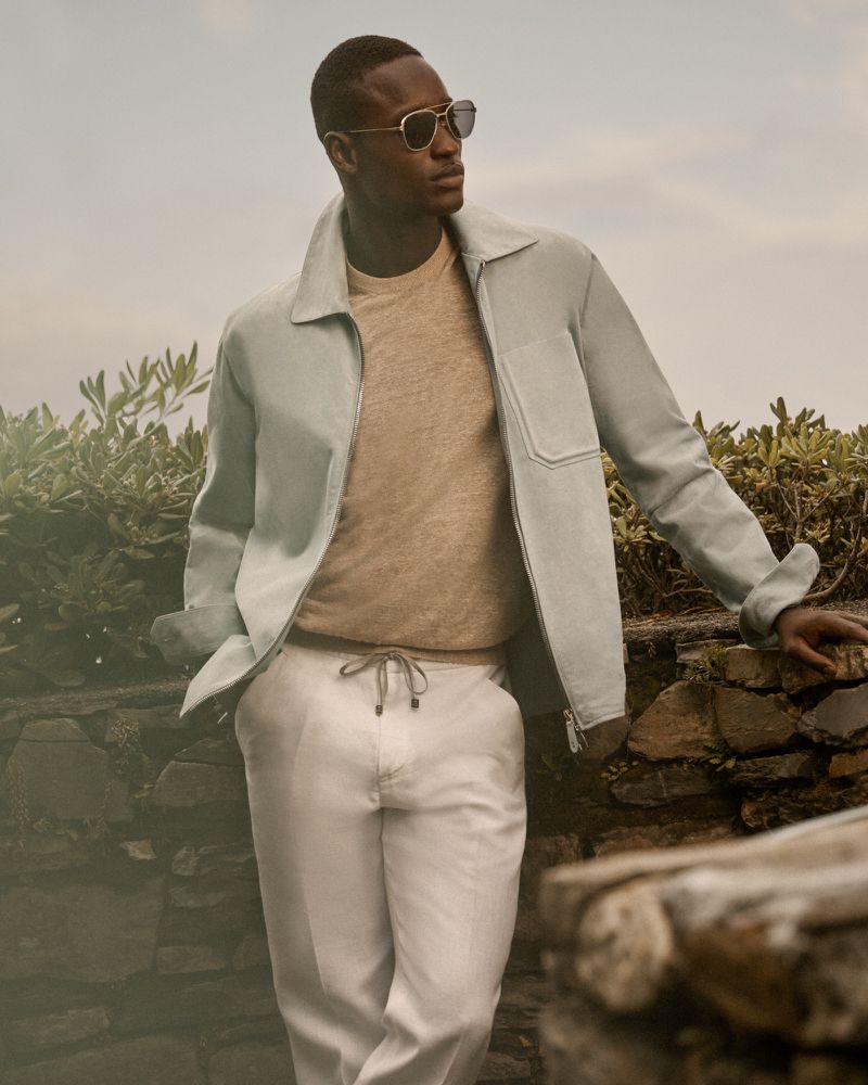 An African American man outside poses wearing sunglasses, a light blue jacket, light brown sweater, and light-colored trousers.