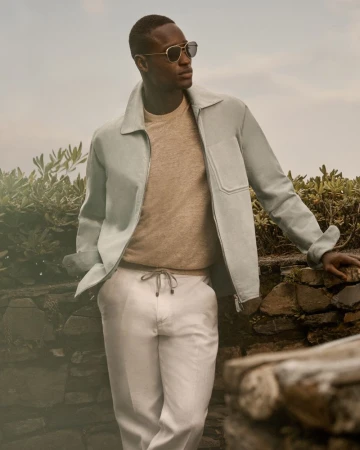 An African American man outside poses wearing sunglasses, a light blue jacket, light brown sweater, and light-colored trousers.