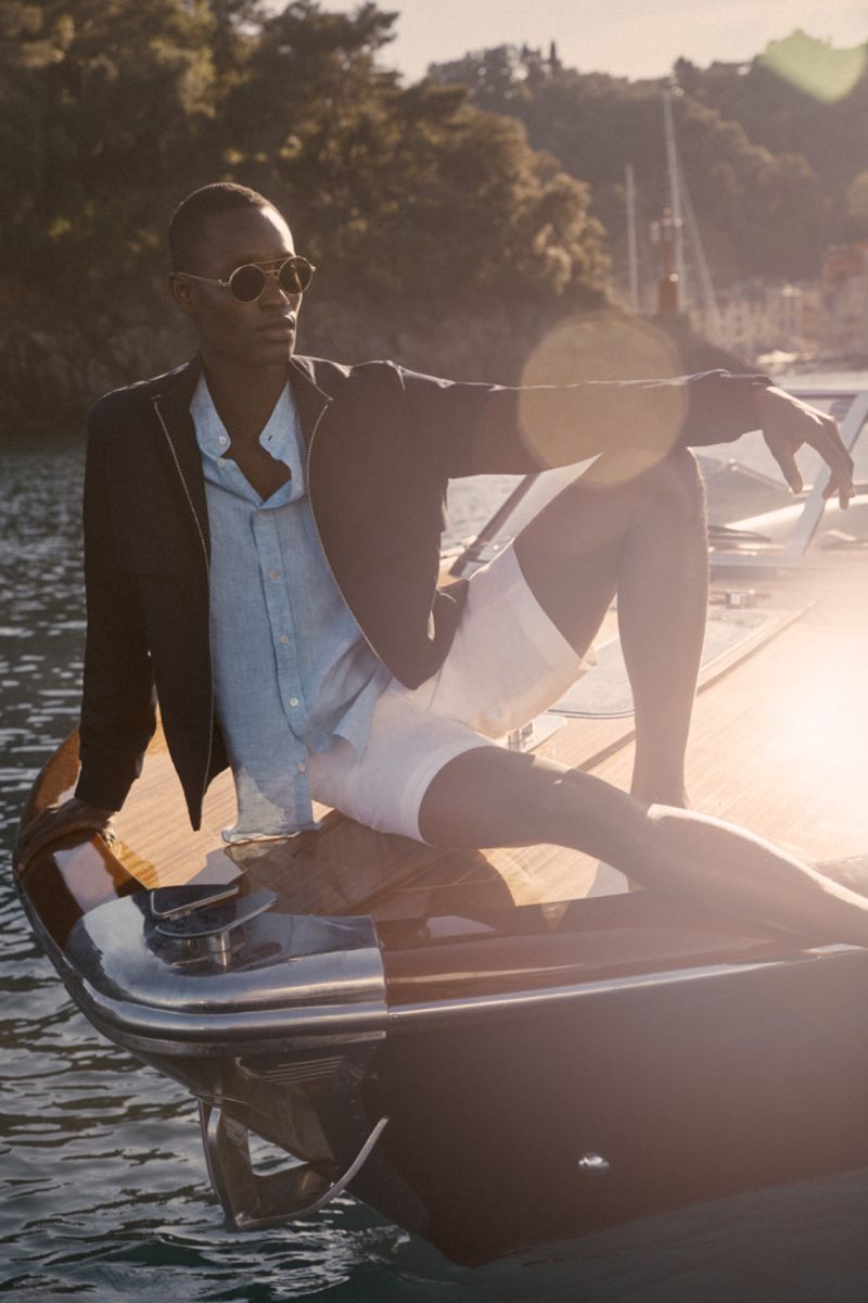 An adult man is lounging on a boat in nautical clothing, sunglasses, and jacket with the ocean and greenery in the background.