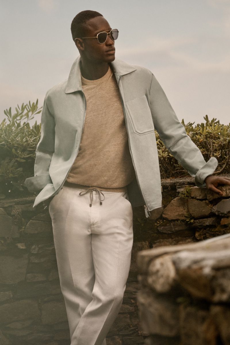 Dark-skinned adult man wearing sunglasses, jacket, and pants poses outside on a stone structure with a neutral facial expression.