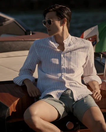 Stylish man in sunglasses and striped shirt seated on a boat with an Italian flag, enjoying a sunny day on the water.