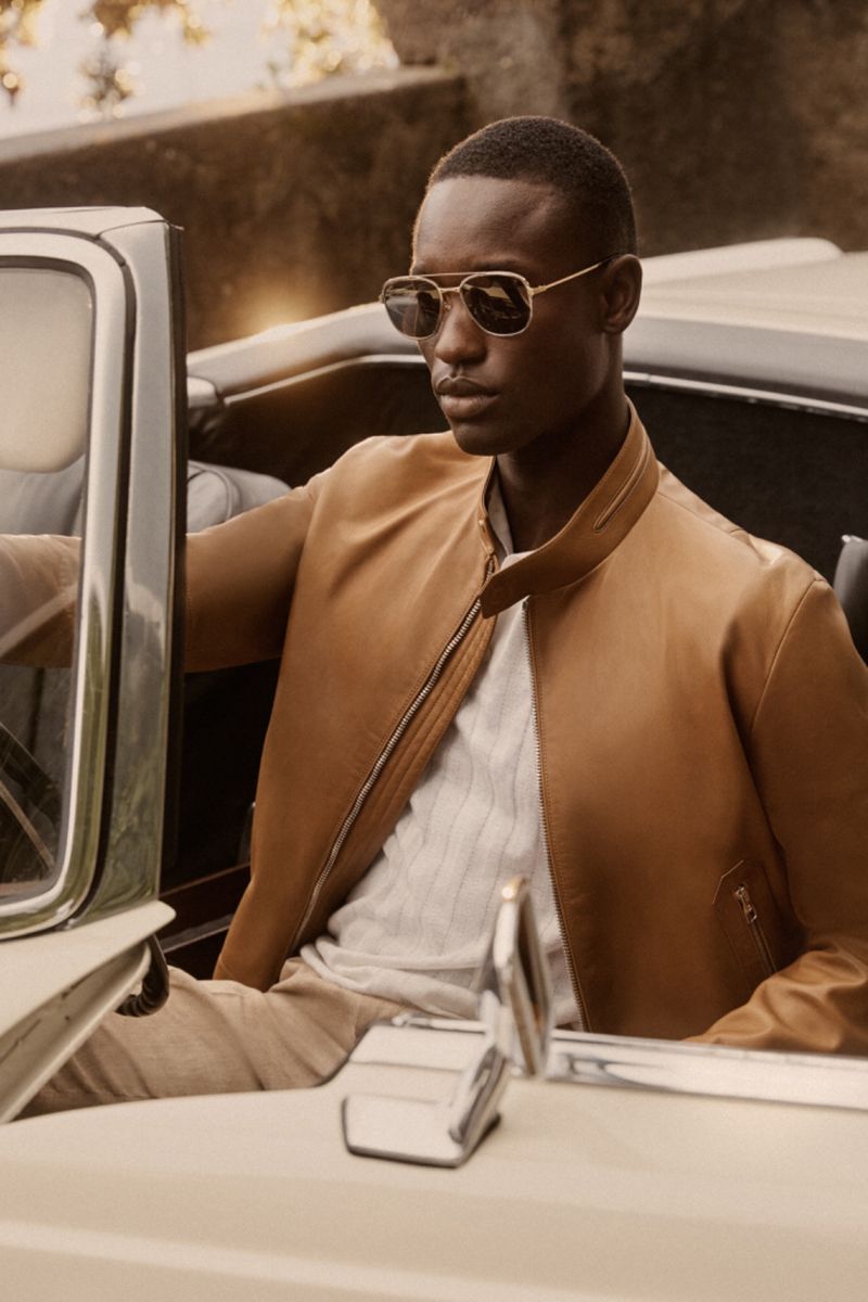 Dark-skinned male model wearing sunglasses and a tan bomber jacket in a classic car. Fashion, lifestyle, and automotive.