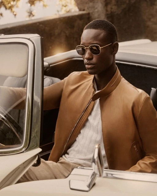 An African American man in sunglasses and a tan jacket sits in a vintage convertible with a cream interior and background foliage.