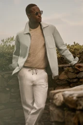 Charles Oduro poses outdoors wearing a light jacket, beige crewneck, sunglasses, and white drawstring trousers by a stone wall.