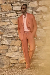Black model wearing an orange double-breasted suit and sunglasses standing against a rustic stone wall