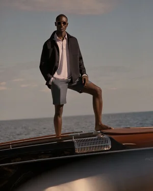 Model standing on the deck of a boat wearing a dark jacket, light polo, and shorts against an ocean backdrop.