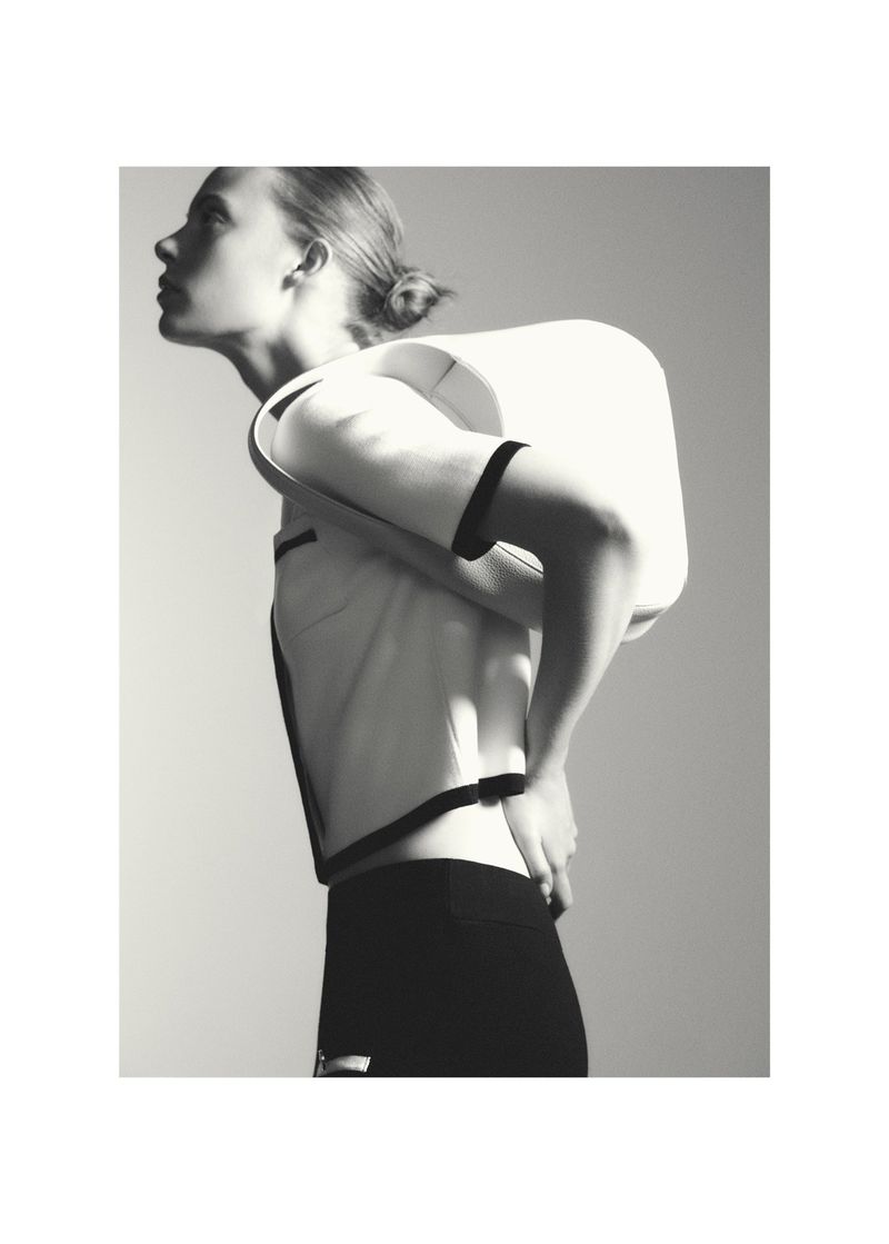 Monochrome fashion shot: woman with hair in bun poses in white short-sleeve top with dark trim and black pants against a light background.