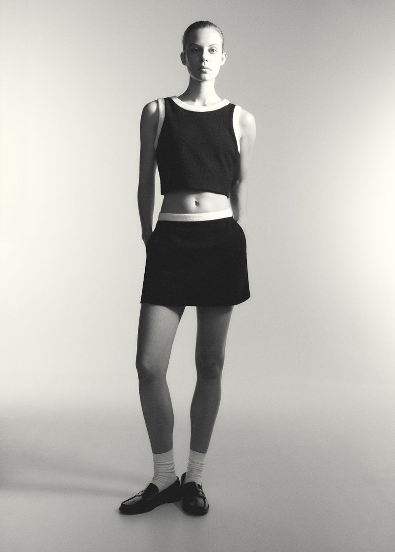 Full-body shot of a young adult woman in a black and white two-piece outfit standing in a bright studio, complemented by matching black shoes.