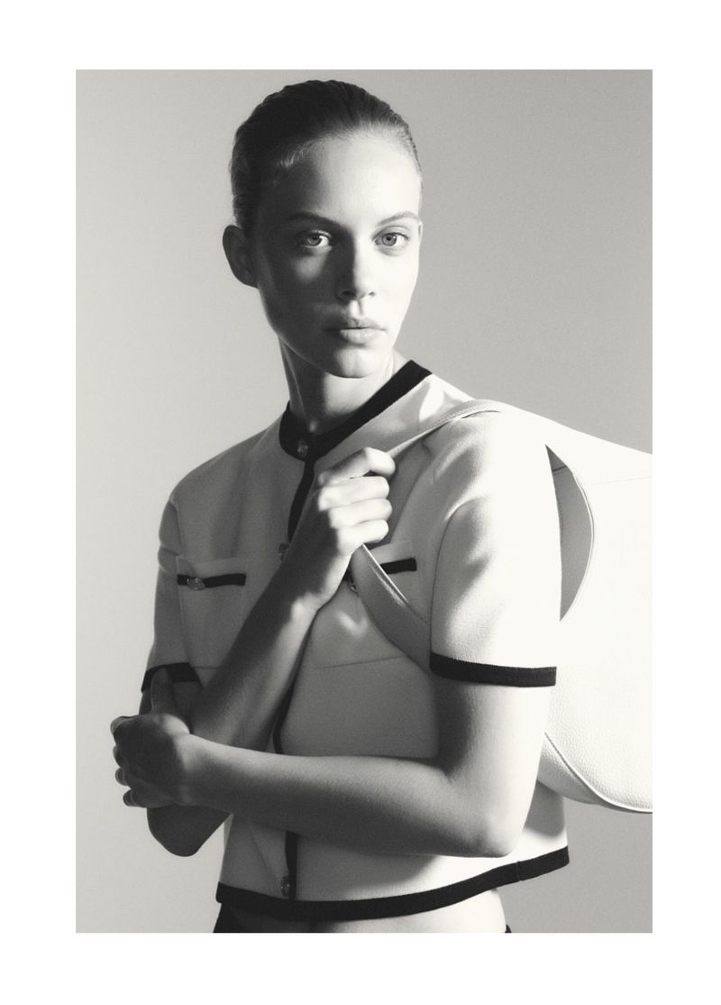 Monochrome portrait of a female fashion model in a stylish white top with a white purse on a plain light background.