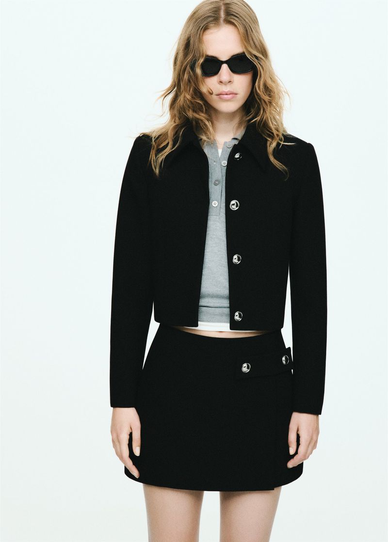 Fashion shot of a blonde woman in a black jacket and skirt with sunglasses against a white backdrop in a studio setting. 