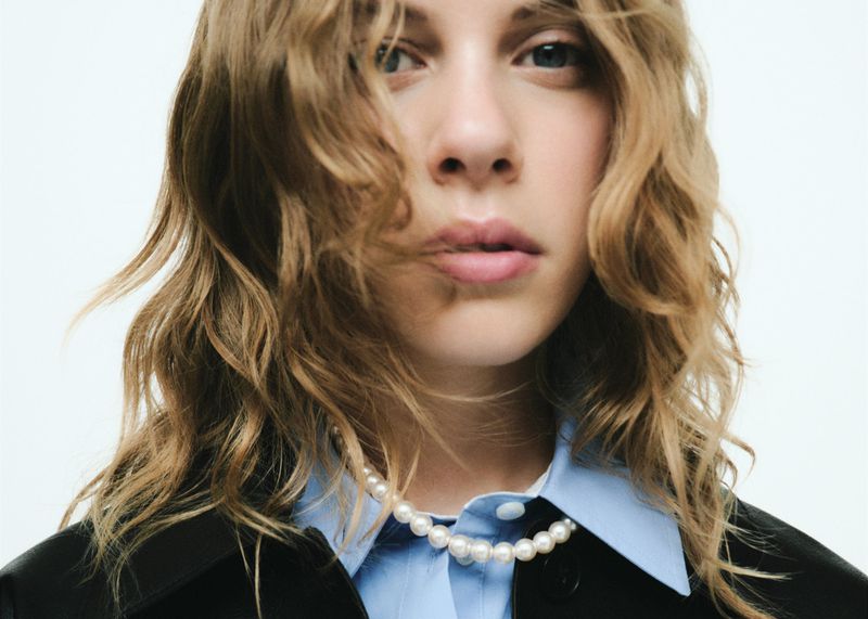 Close-up portrait of a blonde woman wearing a black jacket and a pearl necklace against a white background in a fashion setting.