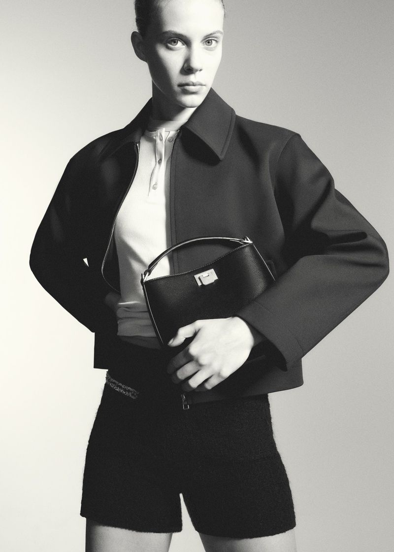 Monochrome studio shot of a model showcasing a black leather handbag with a silver clasp, complemented by a dark jacket and shorts.