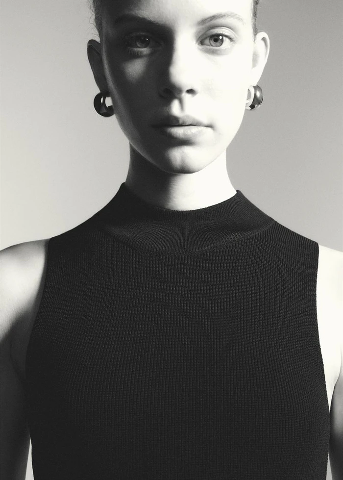 Close-up portrait of a woman wearing a black sleeveless top and hoop earrings against a neutral grey background.