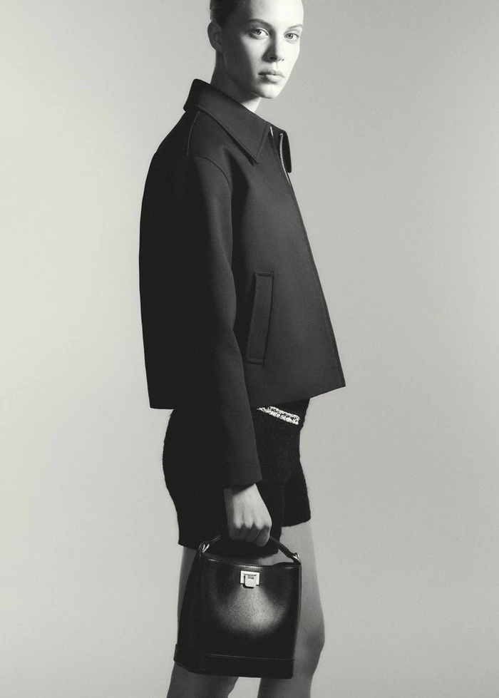 Model wearing dark jacket and holding a black handbag against a grey seamless background