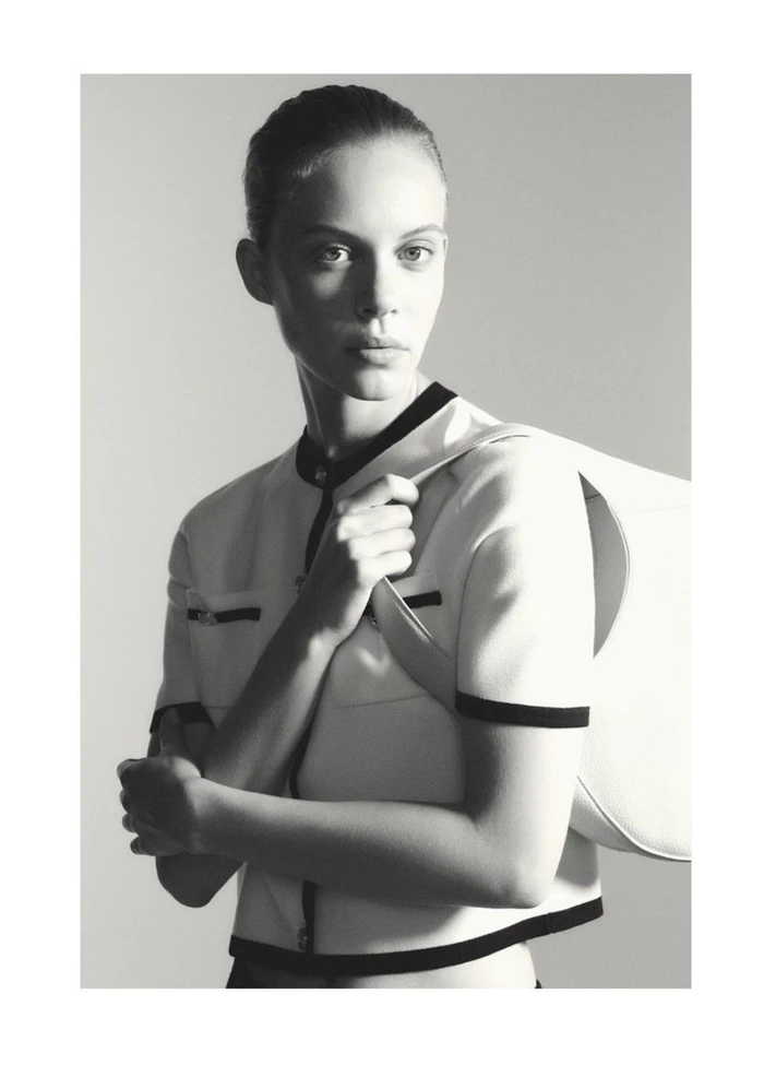 Model Anouk Smits posing in a retro white short-sleeved jacket with black trim and a shoulder bag against a studio backdrop.