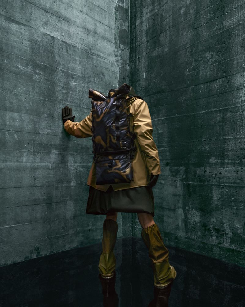 A figure in outdoor fashion, wearing a jacket, skirt, and boots, stands in a concrete corner with a camo backpack against a textural backdrop.