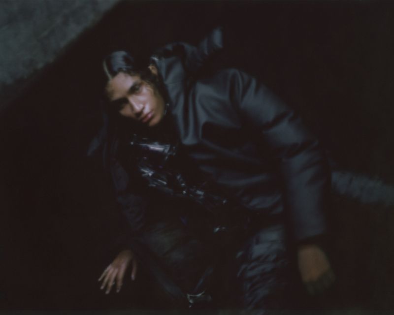 Dark, high-contrast fashion shot of a male model in a hooded jacket, posing on the ground in low light.