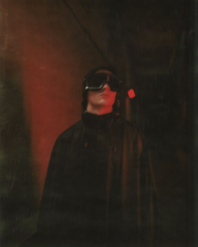 Low angle shot of a person wearing goggles and a black turtleneck in an indoor space illuminated by dim red lighting.