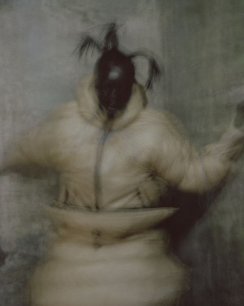 Artistic blurred portrait of a person in a puffy coat with a dark mask and head decorations, set against a textured wall background.